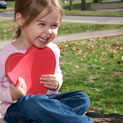 This little girl laughs out loud as I try and take a picture of her. Girl sitting on sidewalk holding a heart
